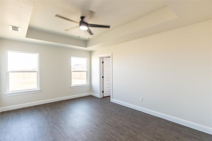 Spacious room featuring tray ceiling with a modern ceiling fan, wood-finish flooring, two large windows, and a panel door