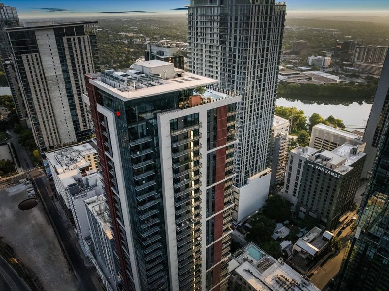 An aerial perspective showcases a sleek high-rise building with a rooftop amenity deck, amidst a vibrant cityscape and a tranquil river.