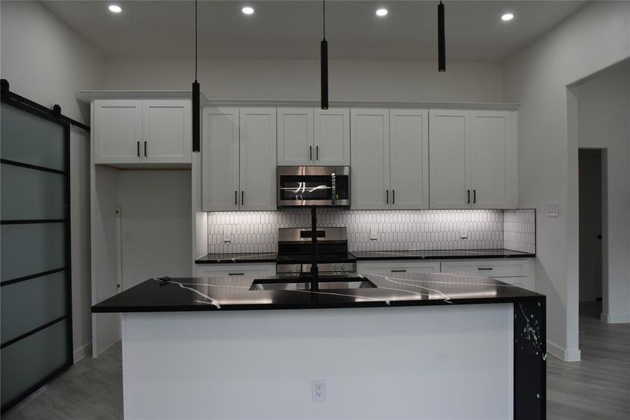 Kitchen featuring white cabinetry, stainless steel appliances, a barn door, an island with sink, and decorative light fixtures