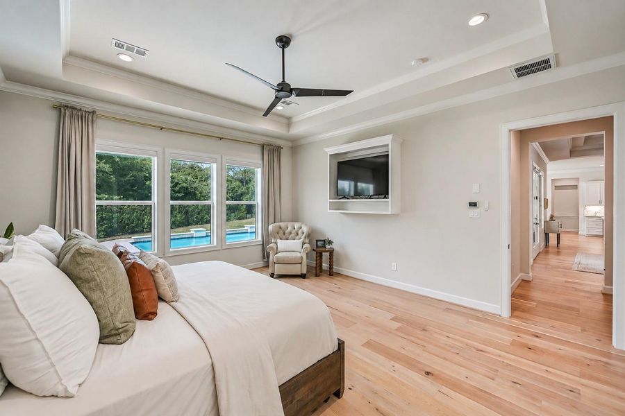 Another view of the primary suite highlights the tray ceiling, abundant natural light and relaxing pool views, plus a built-in media niche and convenient access to the spa-inspired primary bath.