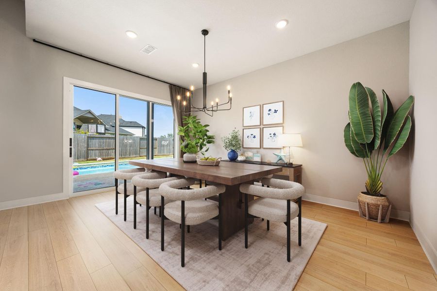 This bright dining area features wide plank wood flooring, neutral walls, and a striking modern light fixture that adds a touch of elegance. Large sliding glass doors open to the backyard and pool, filling the space with natural light and offering seamless indoor-outdoor living.