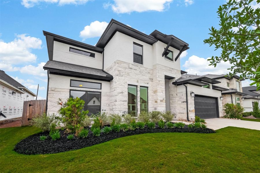 This modern two-story home features a sleek design with a mix of white stone and siding. It includes large windows, a spacious driveway, a two-car garage, and well-manicured landscaping, offering great curb appeal.