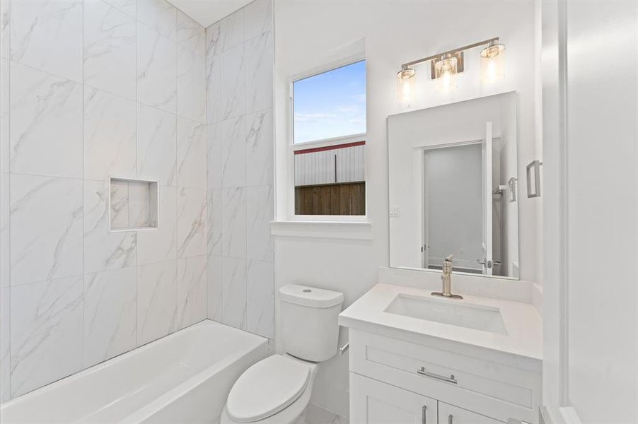 This bright and modern bathroom features a sleek white vanity with a contemporary faucet, a large mirror with stylish lighting, and a clean bathtub with elegant marble-style tile. The room is well-lit by a window, adding to the airy ambiance.