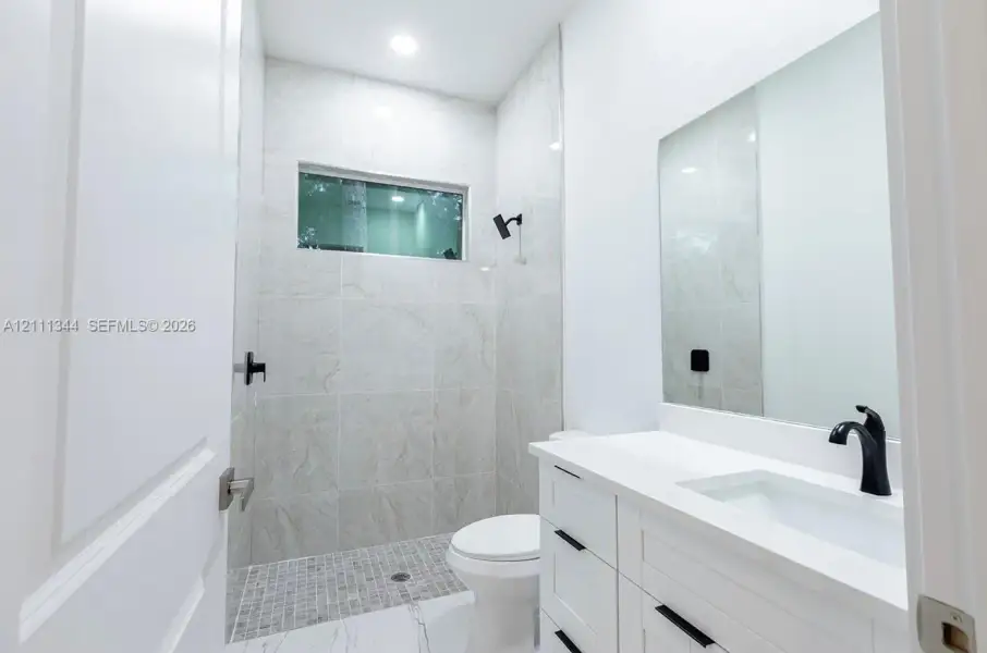 Bathroom featuring a walk-in shower with stone-finish tile surround, a white vanity with a composite countertop, an undermount sink with matte black hardware, and an expansive wall-mounted mirror Bathroom featuring a walk-in shower with stone-finish tile surround, a white vanity with a composite countertop, an undermount sink with matte black hardware, and an expansive wall-mounted mirror
