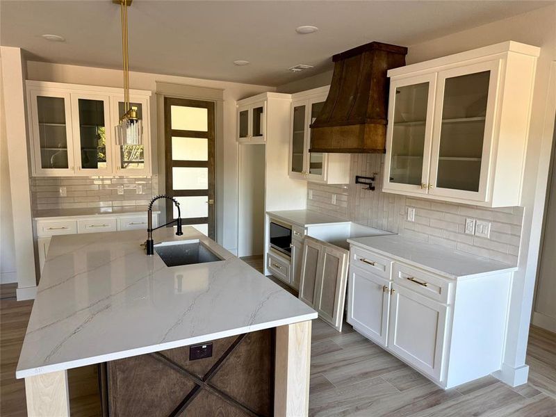 Kitchen with glass insert cabinets, white cabinets, light stone counters, and custom exhaust hood