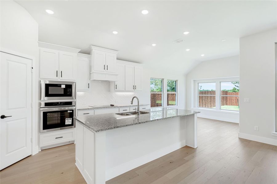 Kitchen with white cabinets, granite island with sink, stainless steel double oven, and light hardwood floors