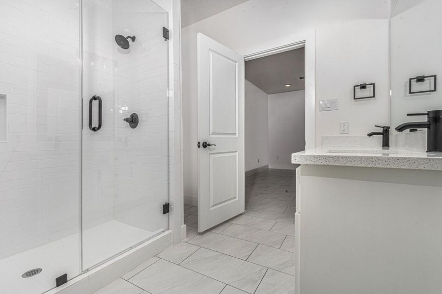 This bathroom features a sleek, modern design with a glass-enclosed shower, white tile flooring, and a spacious vanity with a contemporary black faucet. The space is bright and welcoming, offering a clean and minimalist aesthetic.