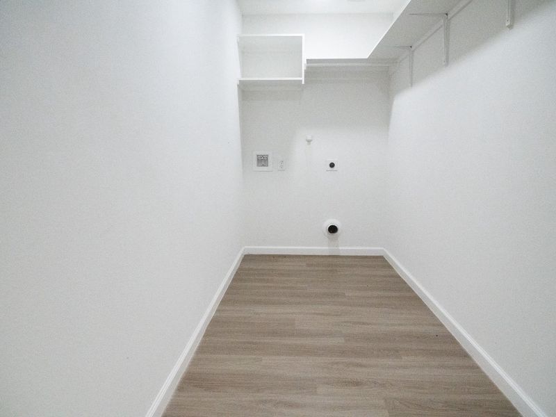 This photo shows a narrow, white-walled utility room with light wood flooring, featuring connections for a washer and dryer and overhead shelving for storage.