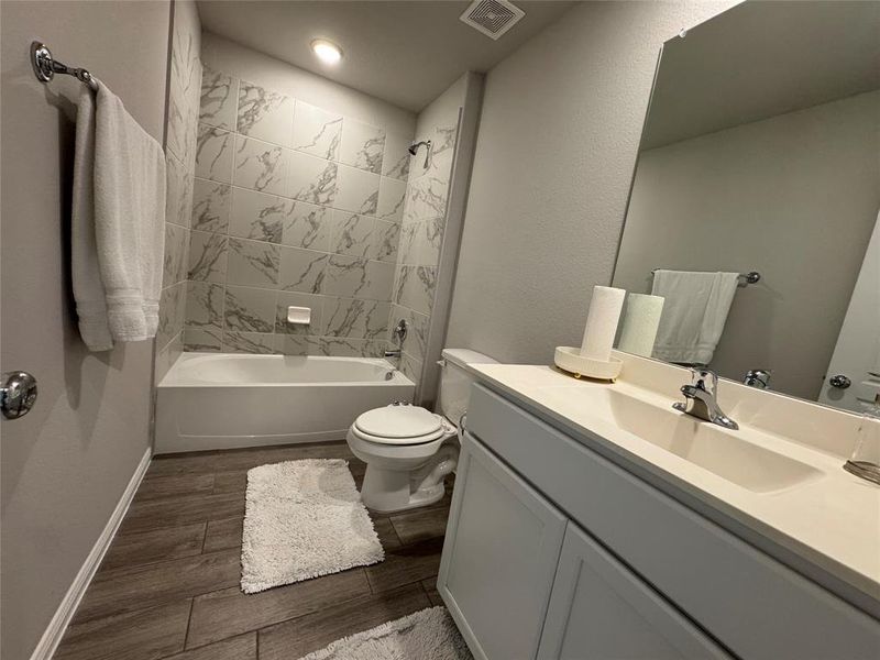 Bathroom featuring a built-in bathtub with a tiled surround, a white vanity with an integrated sink, a large wall mirror, and wood-finish flooring Bathroom featuring a built-in bathtub with a tiled surround, a white vanity with an integrated sink, a large wall mirror, and wood-finish flooring