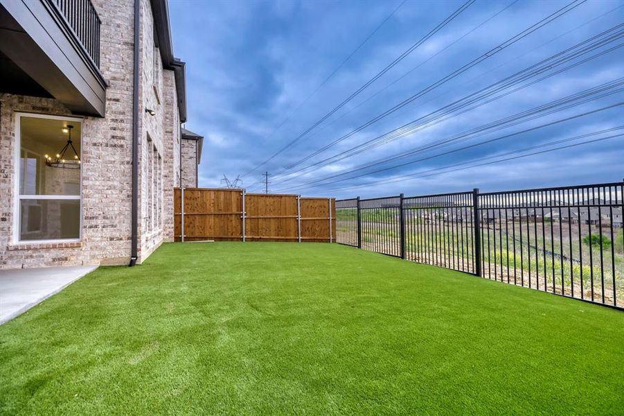 Expansive backyard featuring artificial turf, a concrete patio, a wooden privacy fence, and a black metal fence bordering the property