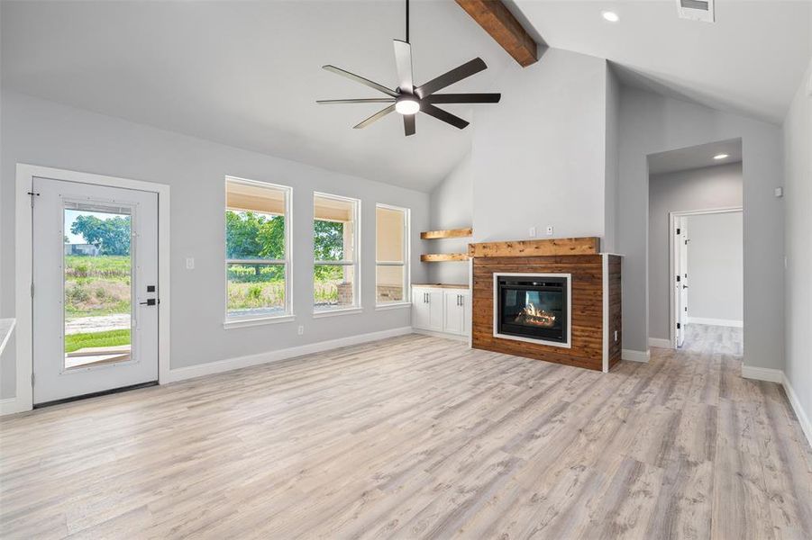 Unfurnished living room featuring ceiling fan, beam ceiling, light wood-style floors, a tiled fireplace, and high vaulted ceiling Unfurnished living room featuring ceiling fan, beam ceiling, light wood-style floors, a tiled fireplace, and high vaulted ceiling