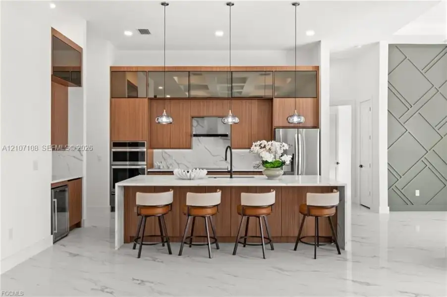 Modern kitchen featuring flat-panel wood cabinetry, a central island with a waterfall countertop, stone-finish backsplash, three glass-pendant lights, and geometric wall molding