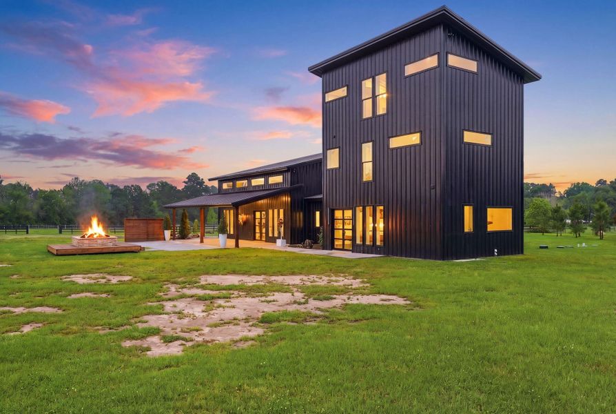 Modern black metal home with large windows and a unique design, set on a spacious grassy lot. Features an outdoor fire pit and scenic views, perfect for enjoying sunsets.