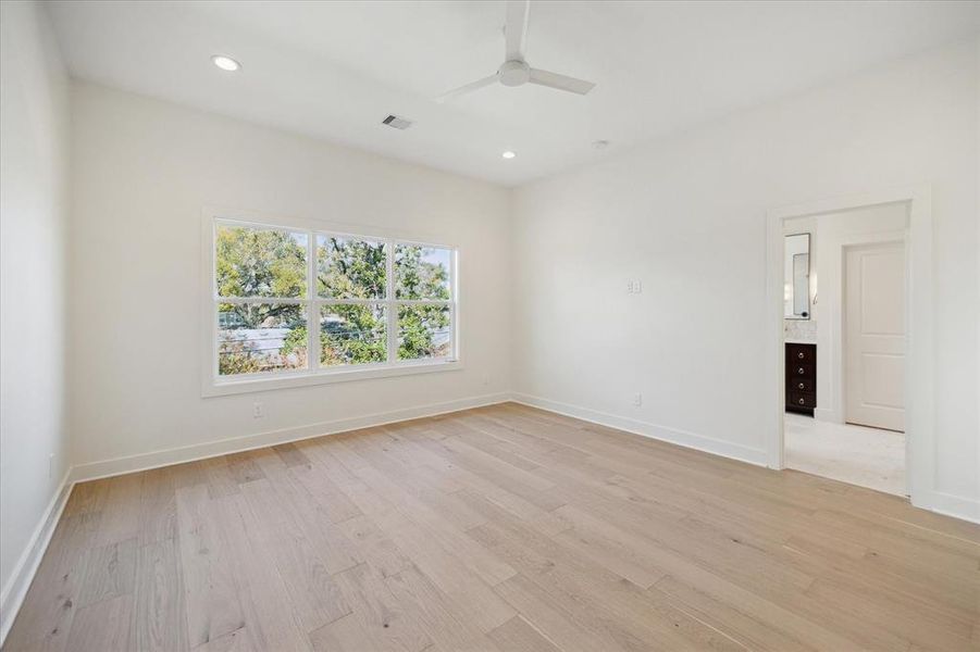 An alternate view of the primary bedroom highlights its spacious layout and abundant natural light. Large windows frame lush treetop views, enhancing the serene atmosphere. The neutral palette and light wood flooring create a versatile backdrop for any design style. A doorway leads seamlessly into the luxurious ensuite bathroom, providing both privacy and convenience.