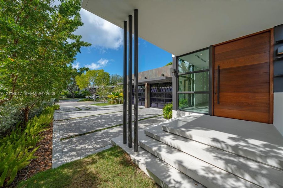 Modern entryway featuring a large wood-finish pivot door, oversized concrete steps, vertical black metal columns, and a paved driveway with grass-infill joints