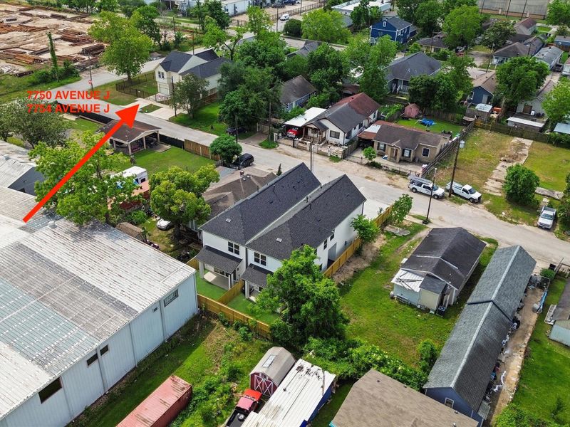 This photo shows a covered carport area attached to a house with beige siding. It opens onto a concrete driveway, enclosed by a wooden fence and a metal gate. There’s some grass and a few trees visible, offering a sense of privacy.