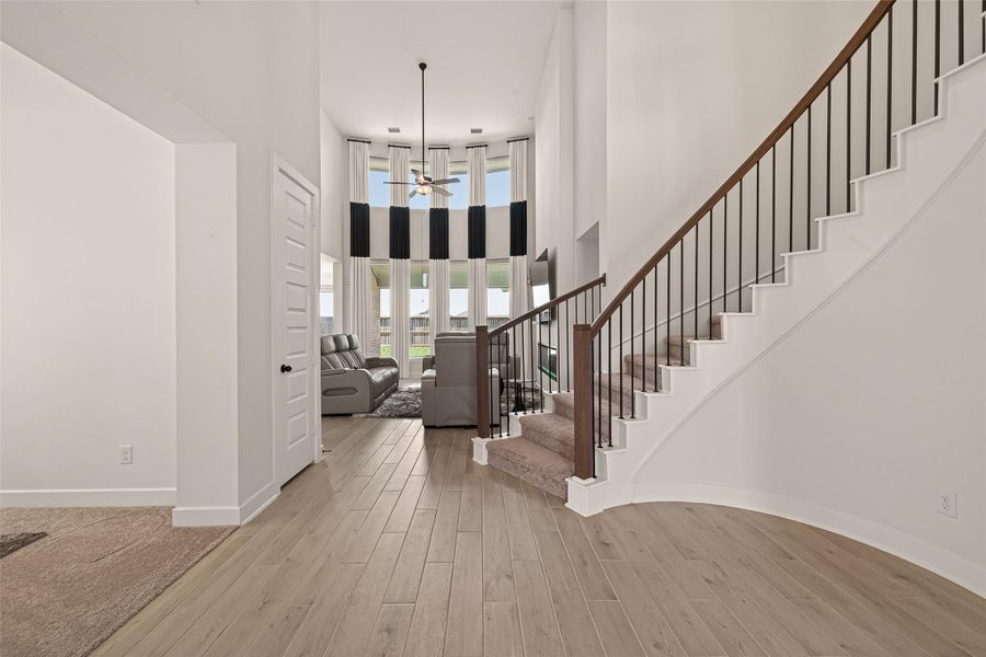 This inviting entryway features a modern staircase with dark wood railings, leading to a spacious, open living area with high ceilings and large windows that allow plenty of natural light. The light wood flooring adds warmth and elegance to the space.