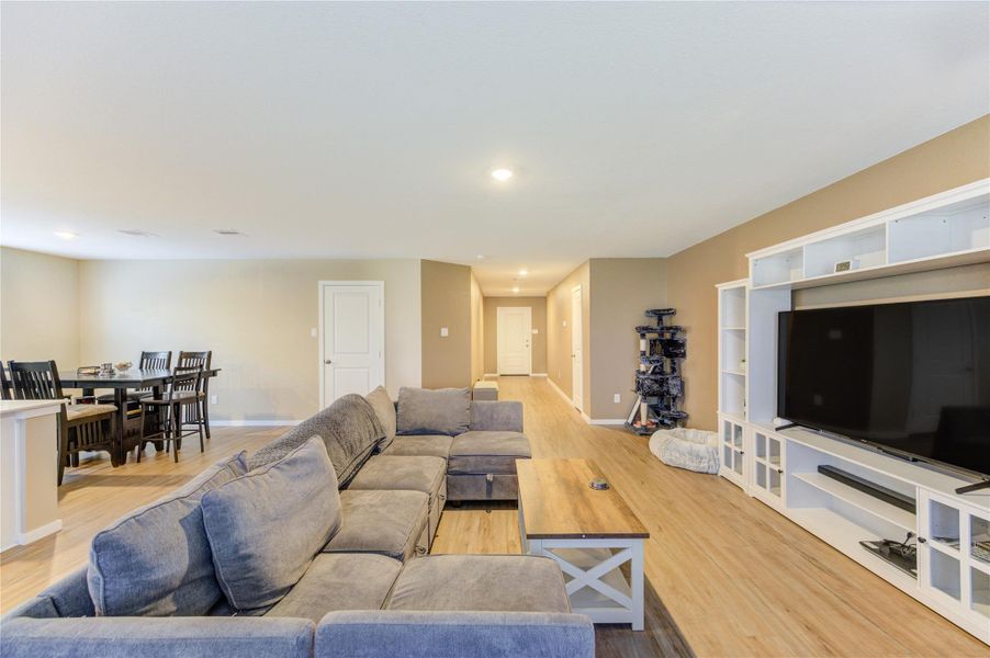 This spacious living area features an open floor plan with light wood flooring, a cozy sectional sofa, a large entertainment center, and a dining area. Ideal for entertaining and family gatherings.