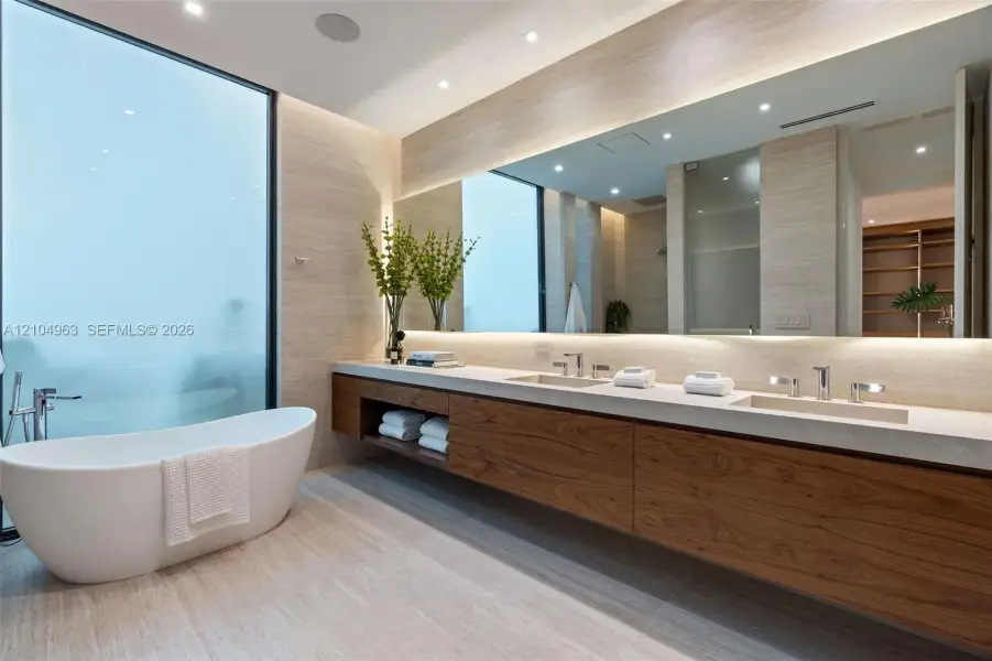 Floating wood-finish vanity with dual integrated sinks, light-toned stone countertop, freestanding soaking tub, expansive wall-to-wall mirror, and recessed ceiling lighting