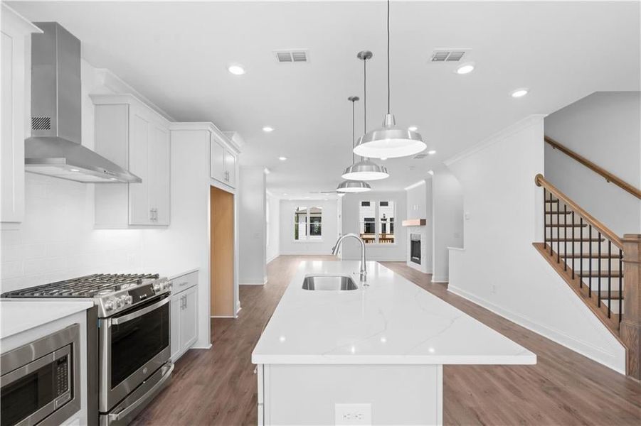 Kitchen featuring a kitchen island with sink, white cabinets, stainless steel appliances, wall chimney exhaust hood, and decorative light fixtures Kitchen featuring a kitchen island with sink, white cabinets, stainless steel appliances, wall chimney exhaust hood, and decorative light fixtures