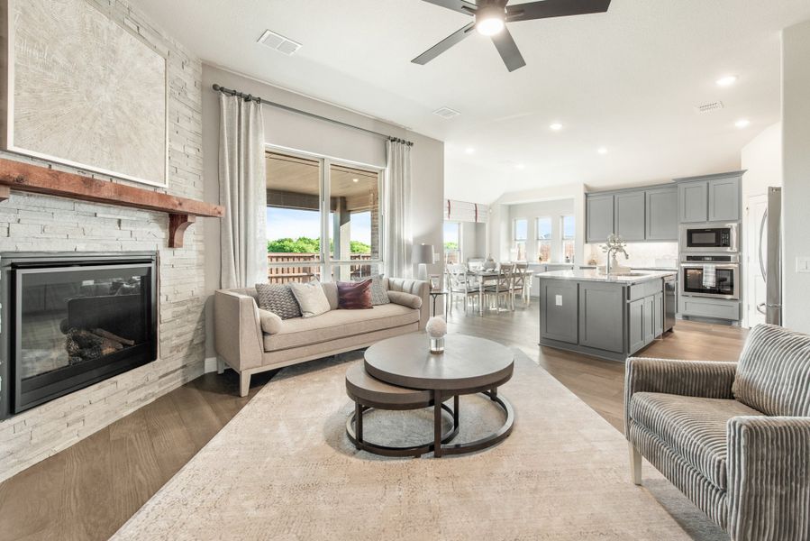 Open-concept living room with stone fireplace and ceiling fan, flowing into kitchen with gray cabinets and island