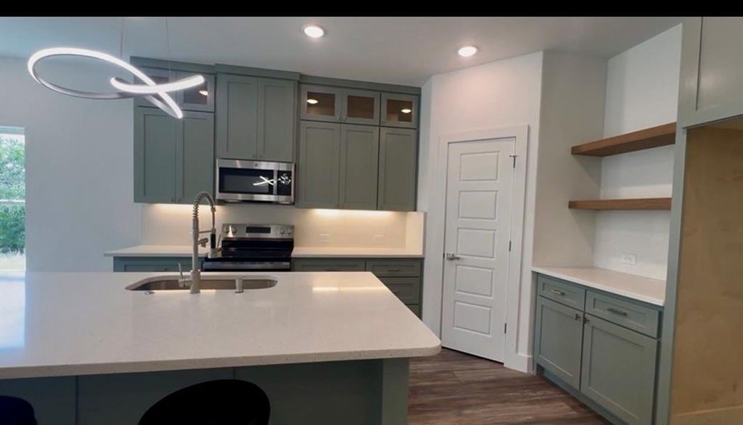 Modern kitchen featuring a large island with an undermount sink, light-colored countertops, sage green cabinetry, stainless steel appliances, and a contemporary pendant light fixture