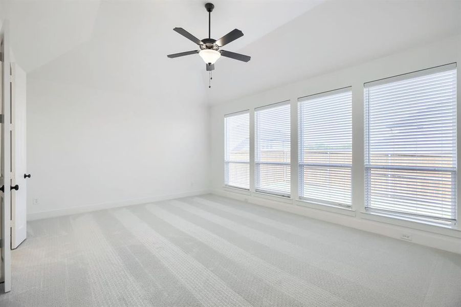 Spacious room featuring vaulted ceilings, a ceiling fan with light fixture, four large windows with horizontal blinds, light-colored carpet flooring, and crisp white walls