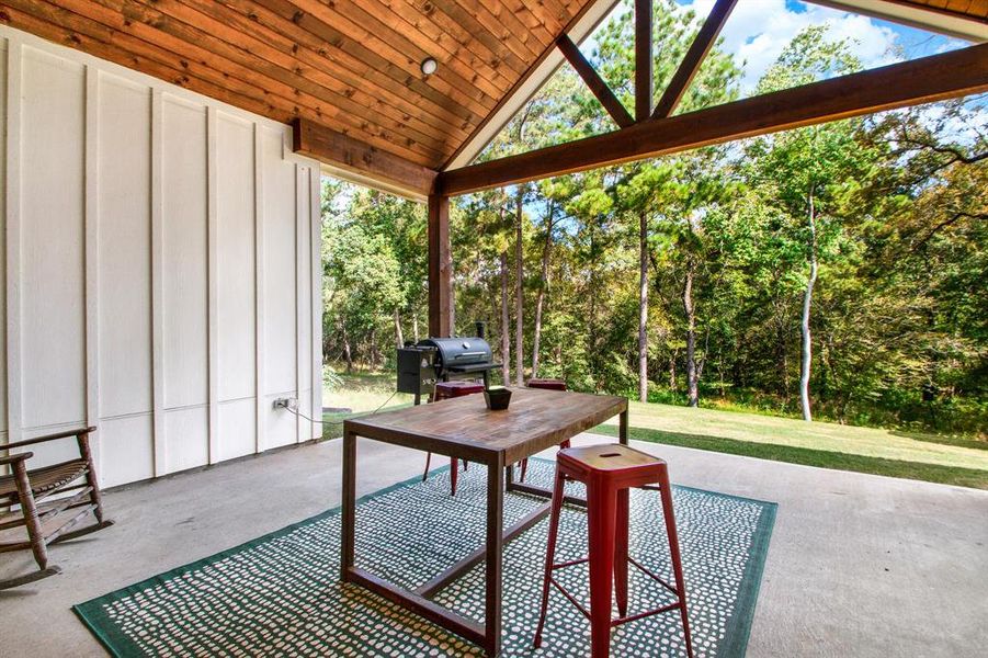 A spacious covered patio area with a high wooden ceiling and large support beams. A large sitting area is a perfect space for a rocking chair along with dining space all overlooking a serene, wooded backyard. Perfect for outdoor dining and relaxation.