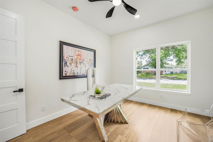 Bright, modern home office with a large window providing natural light, wood flooring, and a sleek marble desk. Ideal for work or study, featuring a stylish ceiling fan and contemporary art on the wall.