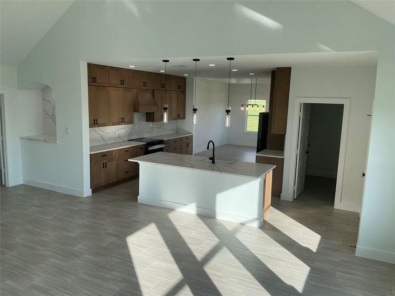 This modern kitchen features sleek wooden cabinets, a large island with a sink, and elegant pendant lighting. Bright natural light floods the space, highlighting the flooring and providing a fresh, airy feel.