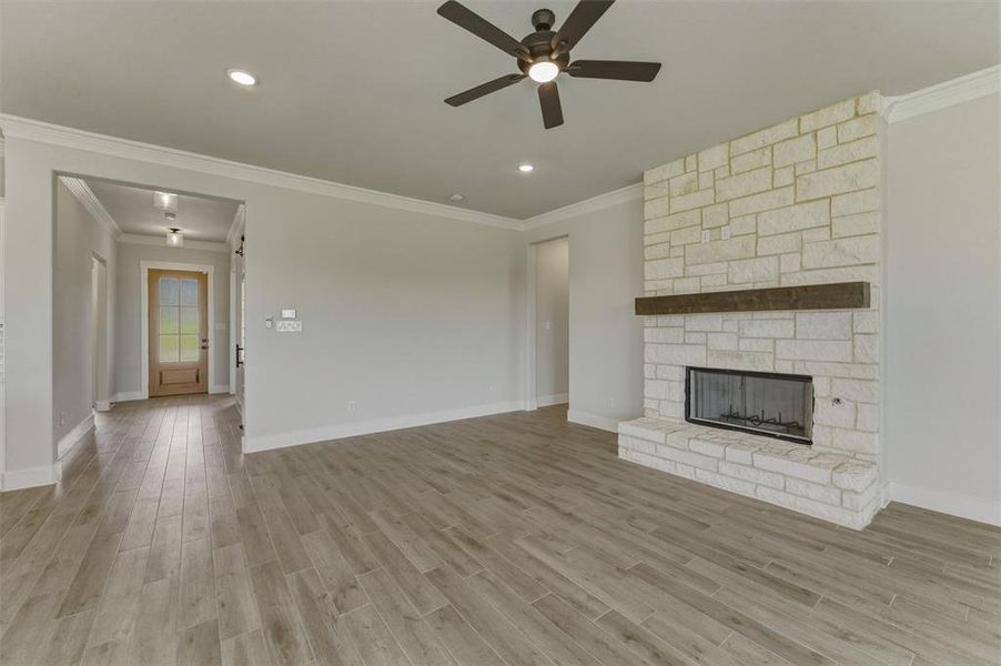Spacious living area featuring a light-toned stone fireplace with a wood mantel, wood-finish flooring, and crown molding Spacious living area featuring a light-toned stone fireplace with a wood mantel, wood-finish flooring, and crown molding