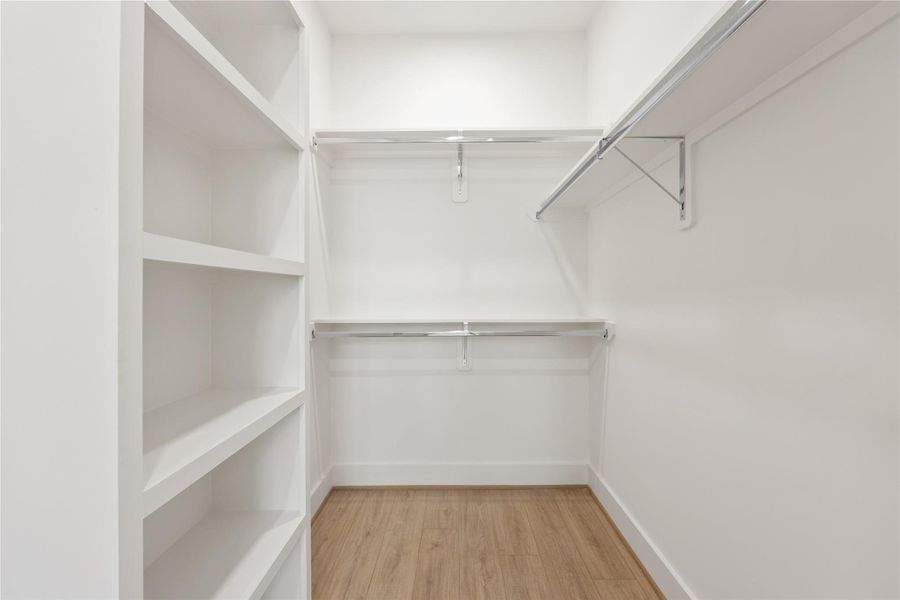 Walk-in closet with built-in shelving, double hanging rods, and warm wood-look flooring provides organized storage and excellent functionality. The clean white finish keeps the space bright and streamlined, offering room for clothing, shoes, accessories, and seasonal storage.