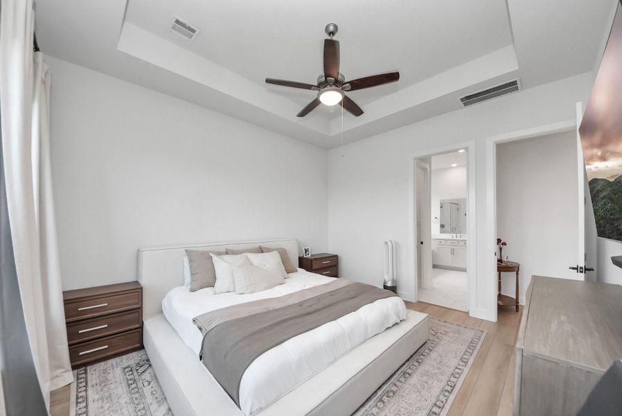 Spacious bedroom with a tray ceiling, modern ceiling fan, and en-suite bathroom, featuring elegant neutral decor and ample natural light.