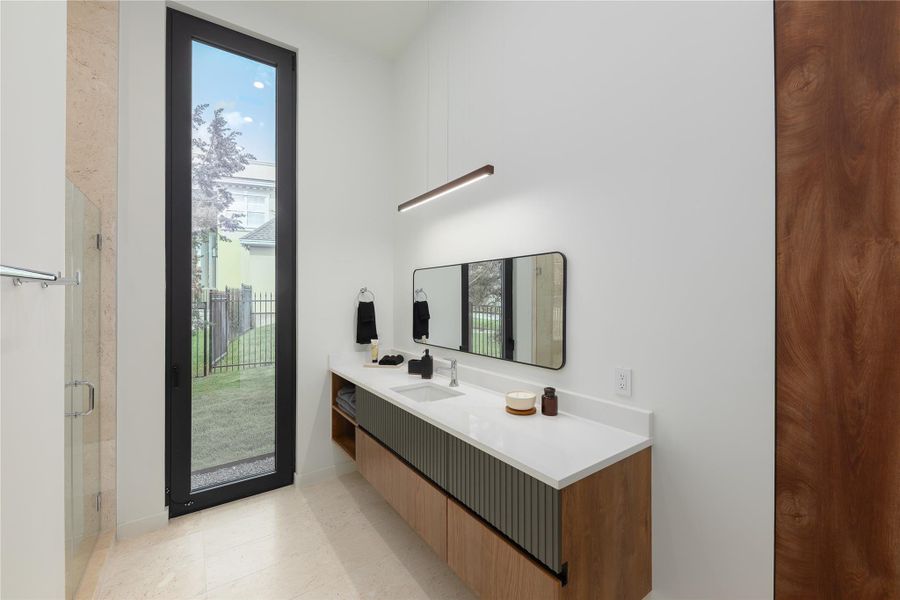 A well-lit bathroom features a long vanity with dual sinks, modern lighting, and a tall window overlooking the yard.