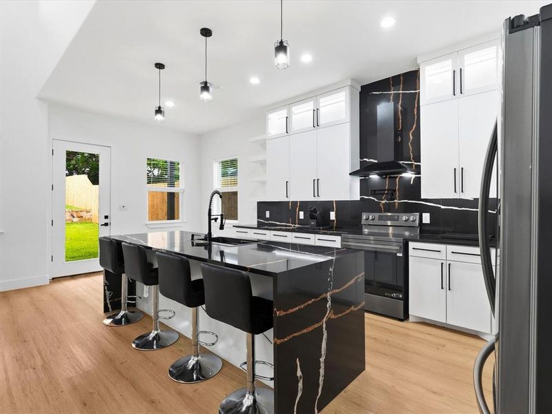 Contemporary kitchen featuring a large island with waterfall countertop, black and gold patterned slab backsplash, white cabinetry with black hardware, stainless steel appliances, and wood-finish flooring