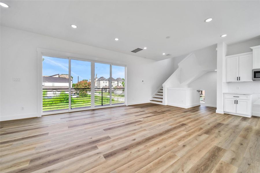 This spacious, open-concept living area features sleek hardwood floors, large sliding glass doors leading to a balcony with neighborhood views, and a modern kitchen with white cabinetry. Natural light enhances the bright, airy feel, complemented by a stylish staircase.