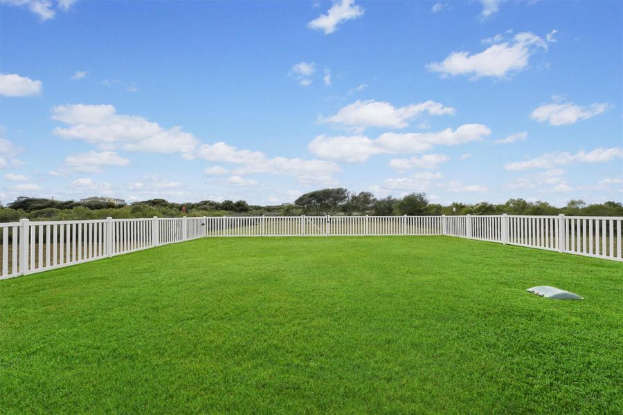 A spacious, well-maintained backyard with with ample room for a pool, hot tub and more. The back yard is enclosed by a weather resistant white fence, offering a clear view of the surrounding natural landscape under a bright, blue sky. fence can even be extended to encompass more of the cleared lot.