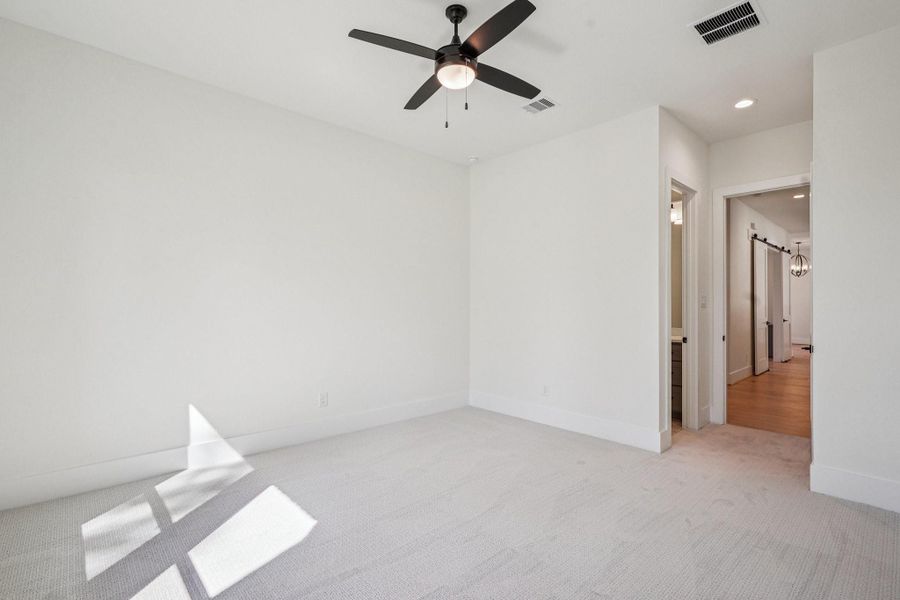 Spacious, bright room with neutral walls and carpet, featuring a ceiling fan. The room leads to a hallway with wooden flooring and a bathroom. Ideal for a bedroom or home office.