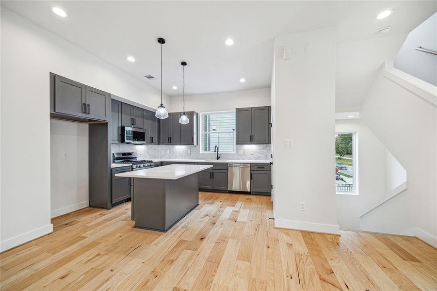 This modern kitchen features sleek gray cabinets, stainless steel appliances, and a center island with pendant lighting. The open layout is complemented by light hardwood floors and ample natural light from large windows.