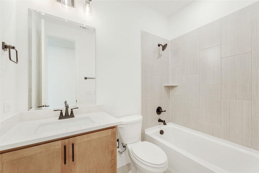 Bathroom with wood vanity, white countertop, toilet, and tiled shower-tub combo with dark bronze fixtures
