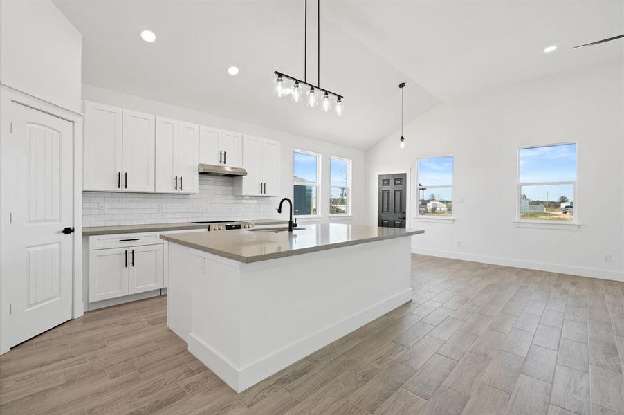 This is a bright, modern kitchen with white cabinets, stainless steel appliances, and a central island. It features high ceilings, recessed lighting, and wood-look flooring. The space is open and airy with natural light coming in through large windows.