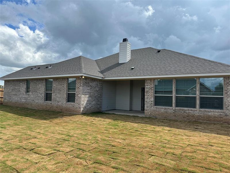 Rear exterior featuring a brick facade, covered patio, multiple windows with blinds, and a shingled roof