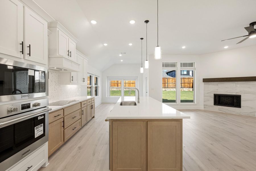 Open kitchen with large island, white quartz countertops, stainless steel appliances, and vaulted ceiling open to living area with fireplace.