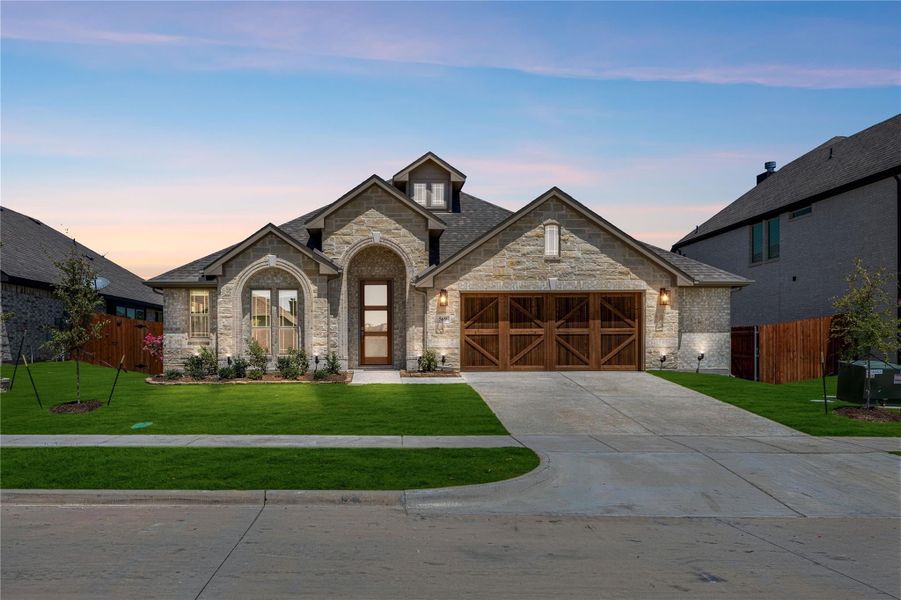 Single-story home with stone exterior, wood garage doors, arched entry, and green front lawn at dusk