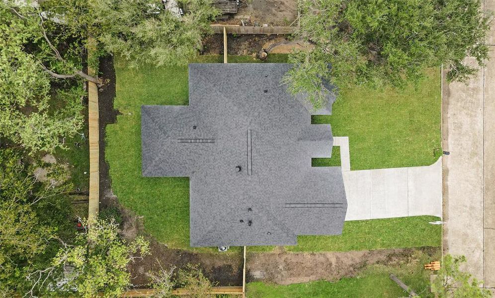 This aerial photo showcases a single-family home with a unique roofline, surrounded by mature trees and a well-maintained lawn. The property includes a driveway leading to an attached garage on a spacious lot, offering privacy and green space.