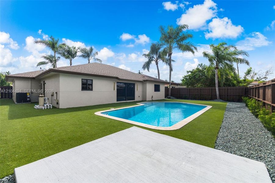 Backyard featuring an in-ground swimming pool, synthetic turf lawn, concrete patio, privacy fencing, and palm trees