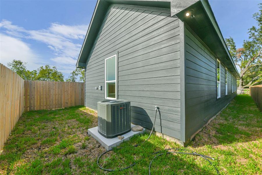 This is an exterior image showing the side of a modern house with gray siding. It features a well-maintained lawn, a wooden privacy fence, and an HVAC unit. The home has multiple windows for natural light.