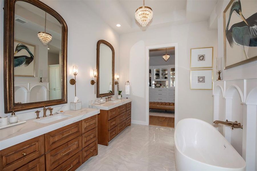 This elegant bathroom features dual sinks with large mirrors, a luxurious freestanding tub, and stylish lighting fixtures. The space is complemented by warm wood cabinetry and a view into a spacious walk-in closet.