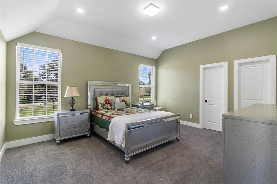 This bedroom is spacious and well-lit, featuring a vaulted ceiling and large windows with views of greenery. It includes a stylish bed, two matching nightstands, and a dresser, creating a modern and cozy atmosphere.