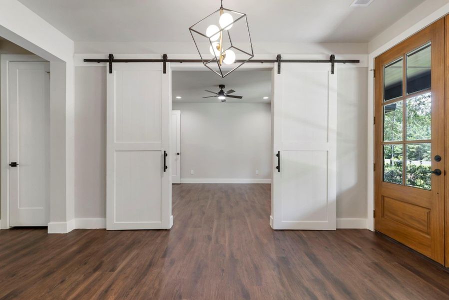 This photo showcases a bright entryway with elegant double wooden doors and glass panels, leading to the study and dining room with plank floors and a large window, creating a welcoming space.